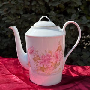 Peut inclure: Théière en porcelaine blanche avec couvercle et anse. La théière présente un motif floral avec des fleurs roses et une bordure dorée. La théière est posée sur une surface rouge.
