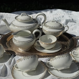 Peut inclure: Service à thé en porcelaine blanche avec bordures dorées, comprenant une théière, un sucrier, une crémière, et plusieurs tasses et soucoupes. L'ensemble est disposé sur un plateau argenté, parfait pour le thé.