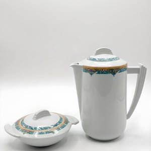 Peut inclure: Une cafetière et un plat de service couverts en céramique blanche. Les deux pièces présentent une bande décorative de motifs floraux turquoise, dorés et roses. La cafetière est haute et élancée avec une anse. Le plat est ovale avec un couvercle et des poignées.