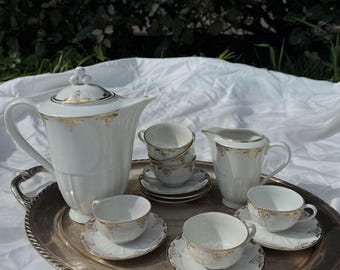 Puede incluir: Un juego de té de porcelana blanca con detalles dorados, que incluye una tetera, una jarra de leche, un azucarero y varias tazas y platillos. El juego está dispuesto sobre una bandeja plateada, perfecto para una merienda.