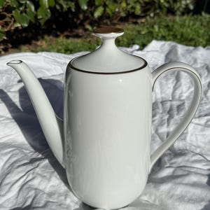 May include: A white ceramic coffee pot with a long spout and a curved handle. The pot has a gold-colored rim and a gold-colored top. The coffee pot is sitting on a white surface.