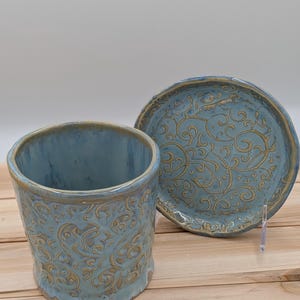 May include: A ceramic cup and saucer set. The cup and saucer are light blue with a detailed embossed pattern and a gold rim. The set is displayed on a light wood surface.