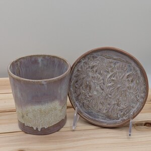 May include: A handmade ceramic planter and matching saucer set. The planter is cylindrical with a lavender and cream glaze. The saucer features a floral pattern. The set is displayed on a wooden surface.