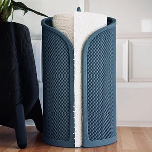 Pode incluir: Um porta-toalhas de papel azul com uma superfície texturizada. O suporte cilíndrico contém um rolo de toalhas de papel brancas. Está sobre uma superfície de madeira, com um vaso de plantas preto ao fundo.