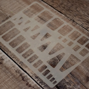 May include: A clear plastic stencil with various shapes, including rectangles, squares, and triangles. The stencil is lying on a wooden surface.