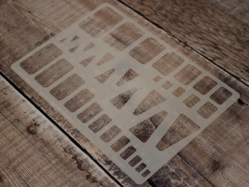 May include: A clear plastic stencil with various shapes, including rectangles, squares, and triangles. The stencil is lying on a wooden surface.