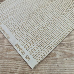 May include: Close-up of a rectangular, off-white bamboo sheet with a pattern of small, evenly spaced, brown ovals. The text "bamboo SM" is visible in the lower left corner, with additional text above it. The sheet is on a light brown wooden surface.