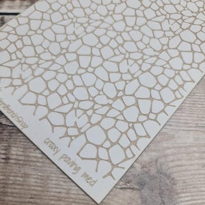 Puede incluir: Un esténcil blanco con un diseño de pavimento loco en color beige. El texto "crazy paving med" está impreso en la esquina inferior derecha. Ideal para proyectos de manualidades.