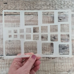 Puede incluir: Una plantilla de plástico blanco con múltiples recortes cuadrados y rectangulares de diferentes tamaños. La plantilla se sostiene en una mano.