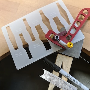 May include: A jewelry-making setup featuring a clear plastic template with various shapes, a red and silver metal tool, a black Sharpie marker, and a silver metal strip with a floral design. The text "The Jewelers Bench" is visible.