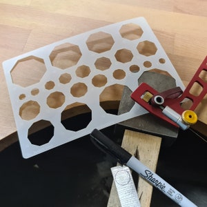 May include: A white plastic stencil with various sized octagon shapes, a red and silver tool, a black Sharpie marker, and a wooden surface.