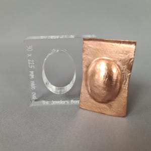Puede incluir: Una herramienta transparente de acrílico para hacer joyas, con un óvalo recortado y el texto "30 x 22.5 mm wide oval" y "The Jeweller's Bench". Junto a ella, una pieza metálica de color cobre con un diseño ovalado.