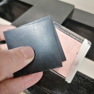 May include: A square black rubber stamp pad with a smooth surface held above a rectangular stamp base. The base features a light pink rectangular rubber surface and a clear acrylic block with text printed along the edge of the transparent frame.