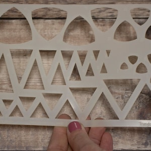 May include: A white plastic stencil with various triangle and teardrop shapes cut out. The stencil is labelled "Triangles" in the bottom right corner.