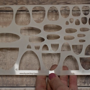 May include: A translucent plastic jewelry stencil template featuring a variety of cut-out shapes including ovals, teardrops, and arches in multiple sizes. The stencil includes numerical markings and the text www.etsy.com/shop/JewelryStencilShop.