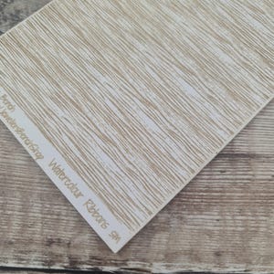May include: A rectangular sheet with a textured pattern of parallel, wavy, light brown lines on a white background. The text "Watercolour Ribbons SM" is printed on a white strip along the edge. The sheet is angled on a wooden surface.