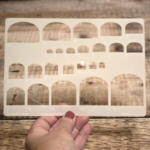 May include: A clear plastic stencil with various arch shapes for crafting. The stencil has measurements in inches, ranging from 8 inches to 45 inches. The text "The Jeweler's Bench" and "Arches I" are printed on the stencil.