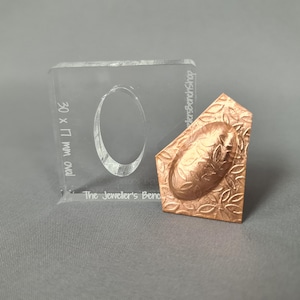 May include: A clear acrylic jewelry making tool with an oval cutout, labeled "30 x 17 mm oval" and "The Jeweller's Bench Shop." Next to it is a copper-colored metal stamp with an oval floral design.