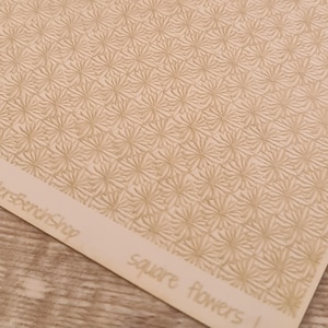 May include: Close-up of a sheet with a repeating pattern of embossed square flowers in a light beige color. The words "square flowers" are printed in a handwritten style on the bottom right corner.
