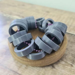 May include: A gray, 3D-printed bracelet composed of circular links. The links are connected by a string of small, pink beads. The bracelet is resting in a small, round, wooden bowl.