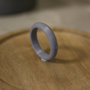 Può includere: Un anello di plastica grigia con una superficie liscia e arrotondata. L'anello è su una superficie di legno.