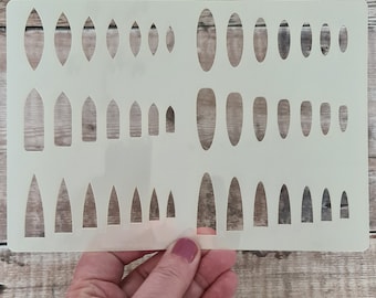 May include: A rectangular, semi-transparent off-white plastic stencil sheet featuring three rows of various nail shape cutouts. The cutouts include almond, square, and stiletto shapes in graduated sizes, arranged in a grid pattern on the sheet.