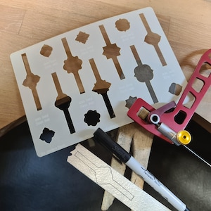May include: A white plastic stencil with various shapes and sizes for jewelry making. The stencil is on a wooden surface with a red metal tool and a black marker pen.