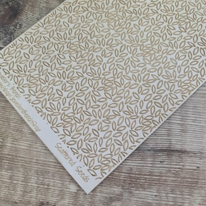 Puede incluir: Una tarjeta blanca con un patrón repetido de formas de hojas de color beige. Las palabras "Scattered Seeds" están impresas en una fuente cursiva en la esquina inferior derecha. La tarjeta está sobre una superficie de madera.