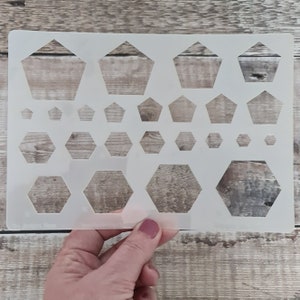 Op de afbeelding: Een witte plastic sjabloon met meerdere maten pentagonen en hexagonen. De sjabloon wordt in een hand gehouden.