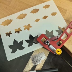 May include: A craft workspace with a white stencil featuring leaf cutouts and a red metal tool. A small, patterned metal piece and a black marker are also visible. The stencil has various leaf shapes for design work.