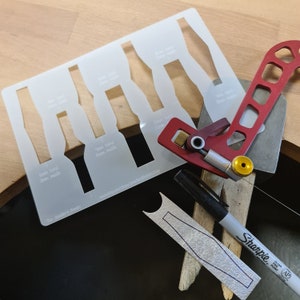 Puede incluir: Una plantilla de plástico blanco con seis formas de banda de anillo de diferentes tamaños, etiquetadas con su tamaño en milímetros. La plantilla se utiliza para crear joyas personalizadas. También se muestra una herramienta de metal rojo y dorado, junto con un marcador negro y una herramienta de metal plateado.