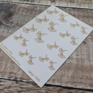 May include: A white card with multiple gold-colored ginkgo leaf motifs. The text "Ginkgo Motifs" is printed in the lower right corner. The card is on a wooden surface.