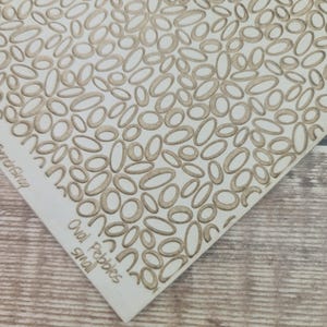 May include: A white rectangular object with a pattern of small, oval-shaped pebbles in a light brown color. The text "Oval Pebbles small" is printed on the object.