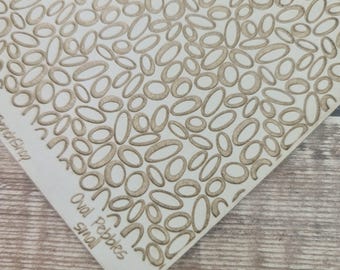 May include: A white square stencil sheet featuring a repeating pattern of small, tan-colored oval pebble shapes. The text "Oval Pebbles small" is printed in tan ink near the bottom left corner of the stencil, which is angled on a wooden surface.