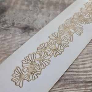Puede incluir: Una tira rectangular blanca con un patrón repetitivo de hojas de ginkgo de color dorado dispuestas en una línea vertical continua. El diseño en oro metálico consiste en delicadas hojas en forma de abanico con detalles lineales finos.