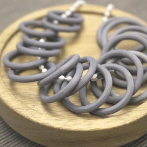 May include: A gray necklace composed of multiple circular rings strung together with white beads. The necklace is displayed on a round, light-colored wooden tray. The rings are a solid, matte gray color.