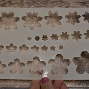 Puede incluir: Plantilla de plástico blanco con múltiples recortes en forma de flor de diferentes tamaños. La plantilla se utiliza para manualidades y la creación de elementos decorativos.