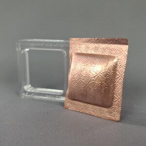 Puede incluir: Un cuadrado de metal en relieve de color cobre con un patrón geométrico, junto a un molde transparente de forma cuadrada. La pieza de metal tiene un centro rectangular en relieve. El fondo es gris liso.