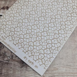 May include: A white sheet with a cracked earth pattern in light brown. The words "dry earth SM" are printed in the bottom right corner. The sheet is angled on a wooden surface.