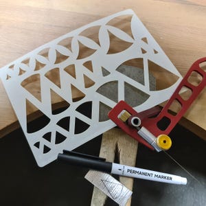 Puede incluir: Una plantilla de plástico blanco con formas geométricas, una herramienta de metal roja y plateada y un marcador permanente negro. La plantilla tiene una variedad de círculos, triángulos y formas curvas. La herramienta tiene un mango y una cuchilla. El marcador está etiquetado como "MARCADOR PERMANENTE".