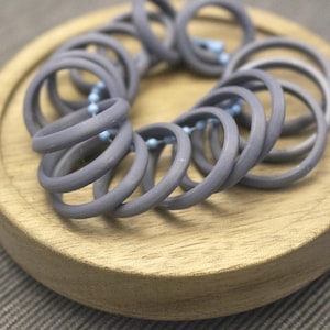 May include: A grey necklace featuring multiple circular rings, connected by small blue beads. The necklace is presented on a round, light-coloured wooden tray, against a neutral backdrop. The rings are evenly spaced, creating a spiral design.