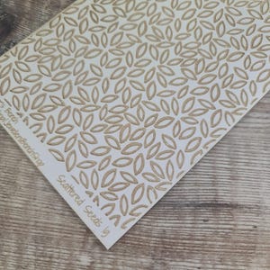 May include: A white card with a repeating pattern of tan leaf shapes. The card has the text "Scattered Seeds" and "JewelersBenchShop" printed on it. The card is on a wooden surface.