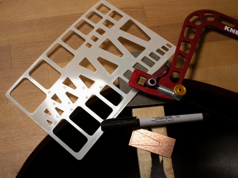 May include: A white plastic template with various shapes and sizes, a red metal tool with a knob, a black Sharpie marker, and a piece of copper with a cut-out shape.