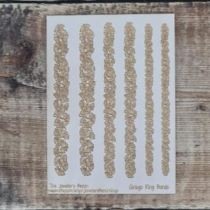 Può includere: Una scheda bianca con sei file verticali di fasce ad anello a forma di foglie di ginkgo dorate. Il testo "Ginkgo Ring Bands" è visibile sul lato destro della scheda. La scheda include anche il testo "The Jeweler's Bench" e un indirizzo web.