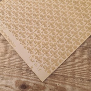 May include: A sheet of cream-coloured paper featuring a repeating pattern of fleur-de-lis motifs. The words "Fleur de Lys" are printed on the corner of the paper. The paper is on a wooden surface.