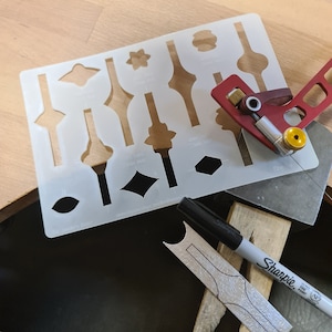 May include: A white plastic stencil with various shapes and sizes cut out. The stencil is used for crafting and woodworking. A red and silver tool is used to cut the shapes out of the stencil. A black Sharpie marker is also visible.