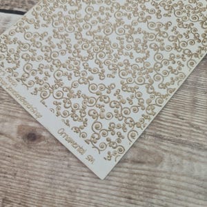 Op de afbeelding: Een crèmekleurige sjabloon met een herhalend patroon van wervelende, sierlijke ontwerpen in een lichtbruine kleur. De woorden "Ornamental SM" staan op de sjabloon.