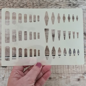 Op de afbeelding: Een witte plastic sjabloon met verschillende uitgesneden vormen. De vormen omvatten cirkels, ovalen, ruiten en driehoeken in verschillende maten. De sjabloon is ontworpen voor knutsel- en doe-het-zelfprojecten.