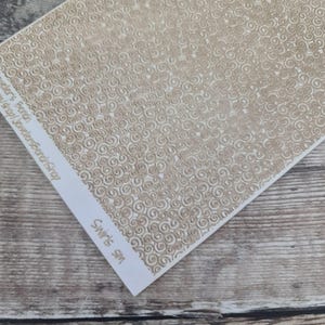 Può includere: Un foglio di carta con un motivo a spirale ripetuto di colore marrone chiaro. La carta ha una superficie strutturata con spirali in rilievo. L'angolo inferiore della carta riporta la scritta "Swirls SM". La carta è su una superficie di legno.