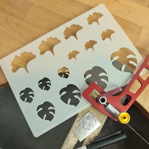 May include: A rectangular white stencil featuring various sizes of ginkgo and monstera leaf cutouts, accompanied by a red metal jeweller's saw frame, a small rectangular metal plate with an etched leaf pattern, and a black marker pen.